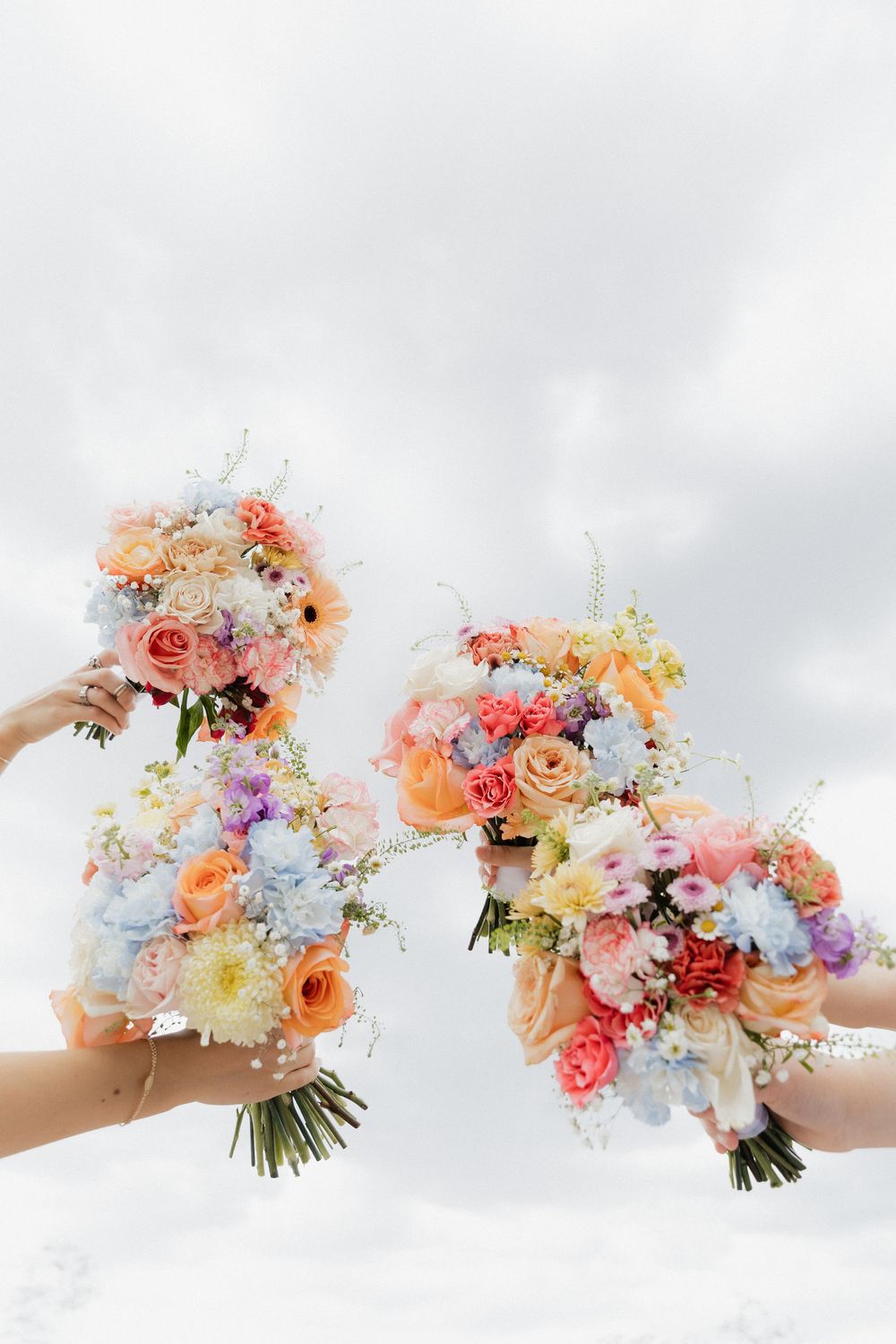 Colorful bridal bouquets with peach roses, blue hydrangeas, and mixed blooms held up against a cloudy sky.