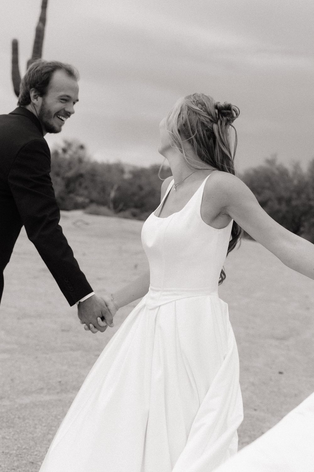 Black and white photo of a smiling bride in white gown and groom in dark suit holding hands on a sandy desert landscape.