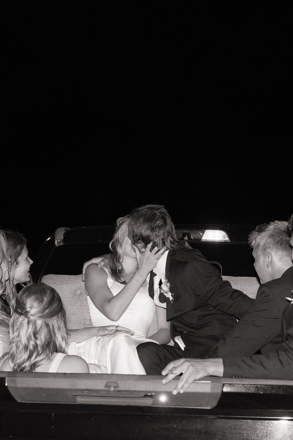 Black and white photo of a bride and groom sharing a kiss in a limousine at night surrounded by wedding guests.