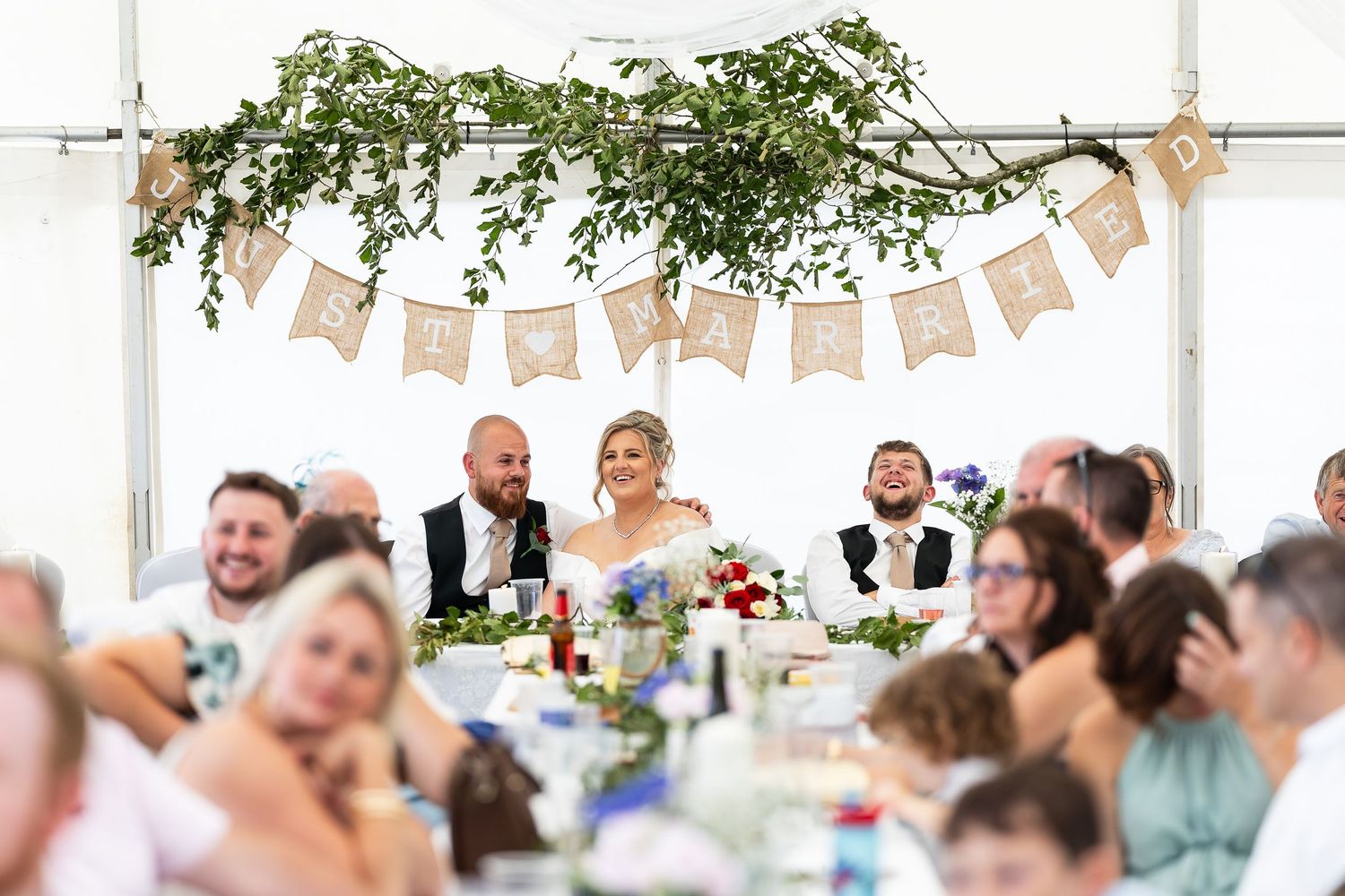 Bride and groom smile at head table during elegant outdoor wedding reception with floral bunting decor.