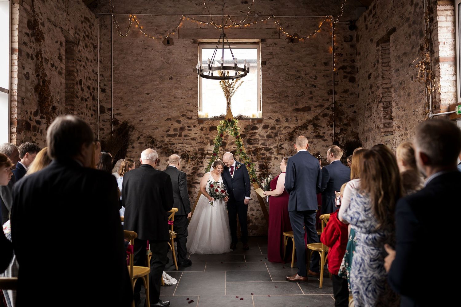 Bride walks down the aisle in a rustic stone barn wedding venue with chandelier and seated guests.