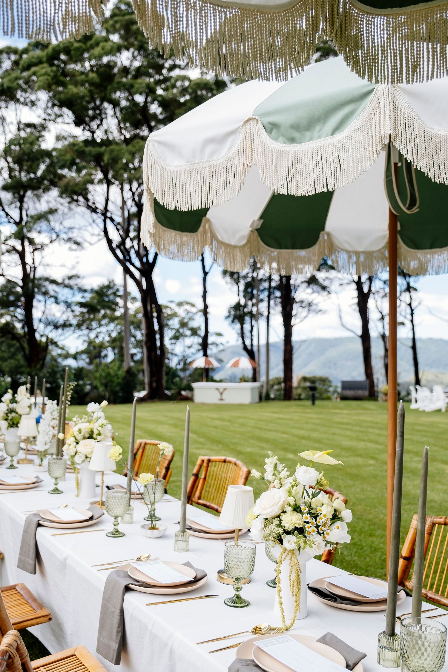 Elegant outdoor wedding reception table set under green and white fringed umbrella with floral arrangements on lush lawn business branding