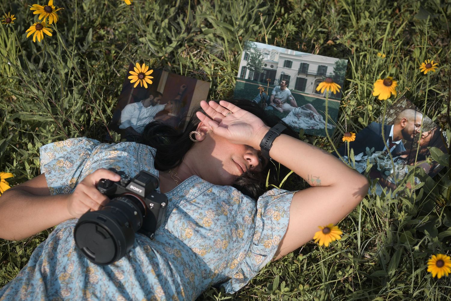 Person in floral dress lying in wildflower field holding camera and photographs outdoors.