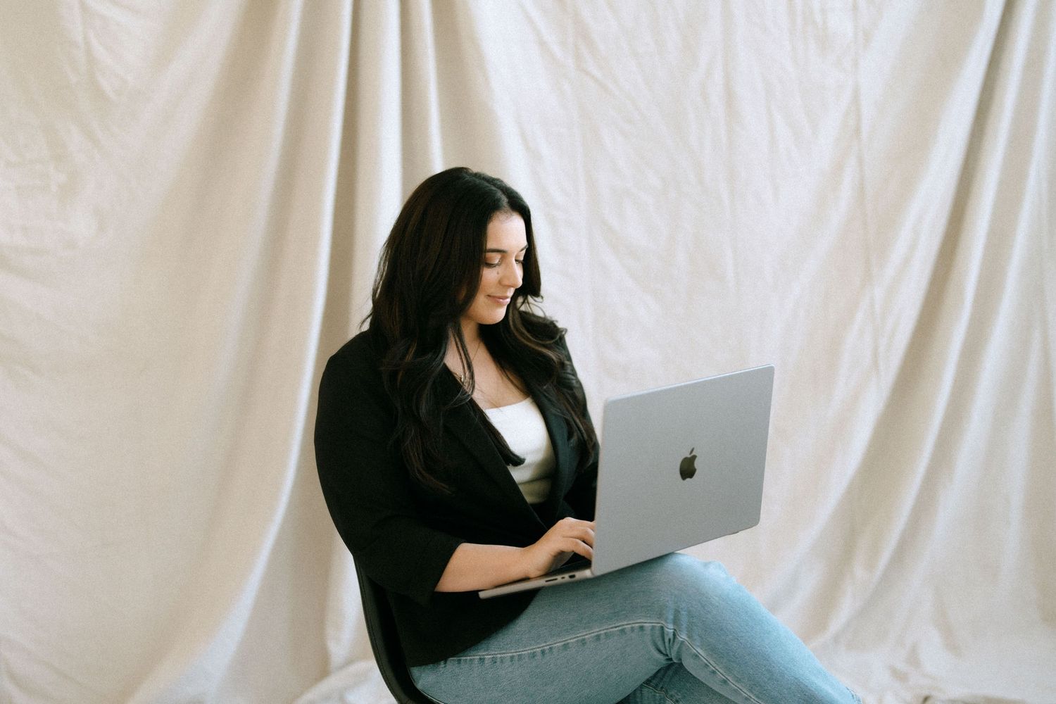 Brunette woman in black blazer working on Apple MacBook laptop against white fabric background.