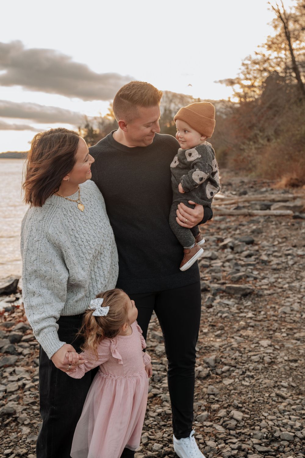 Young family of four sharing a tender moment on rocky lakeshore at golden hour sunset.
