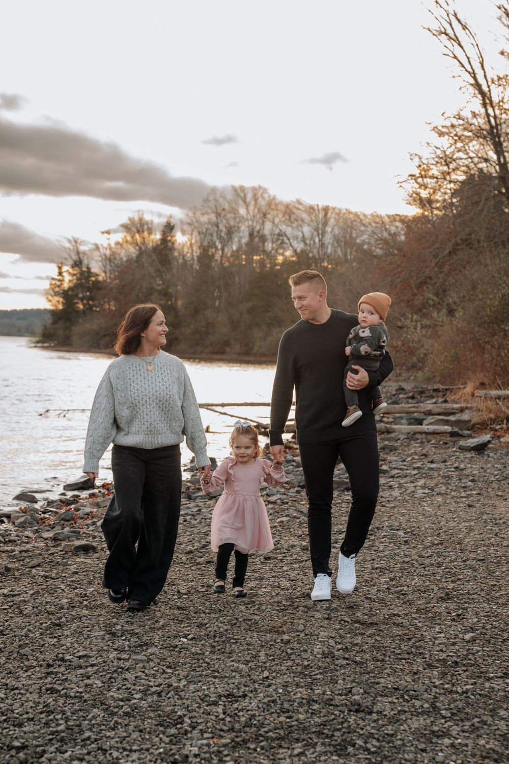 Happy family of four walking hand in hand along rocky lakeshore at autumn sunset.