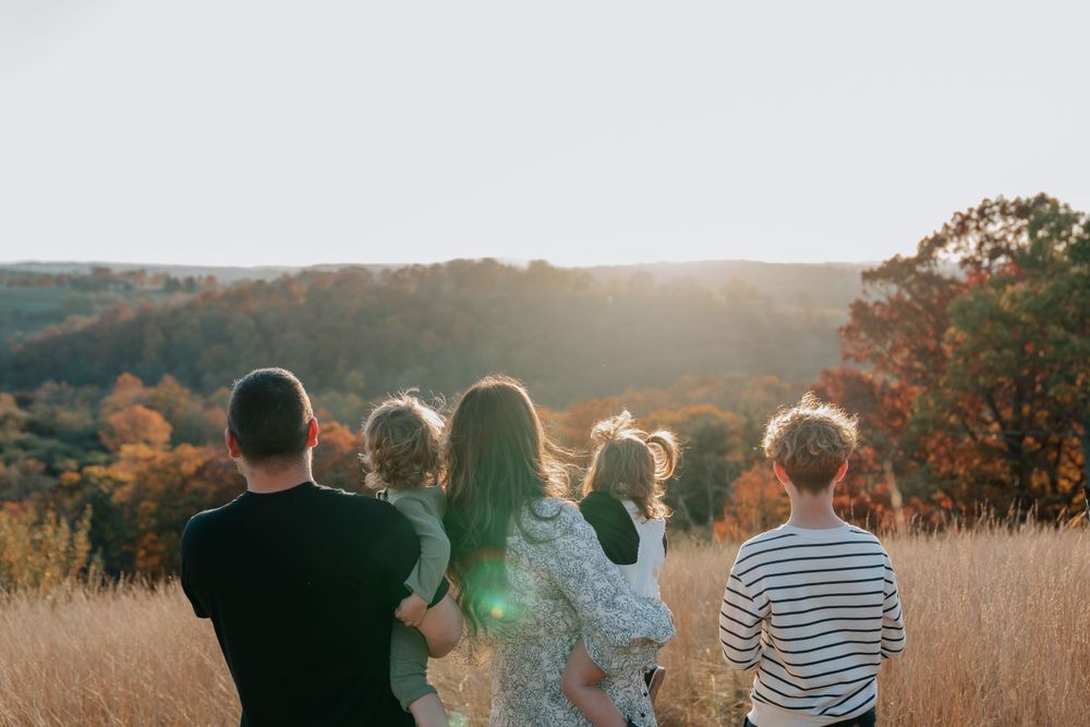 Family of five standing in golden field at sunset, viewed from behind, overlooking autumn forest landscape.