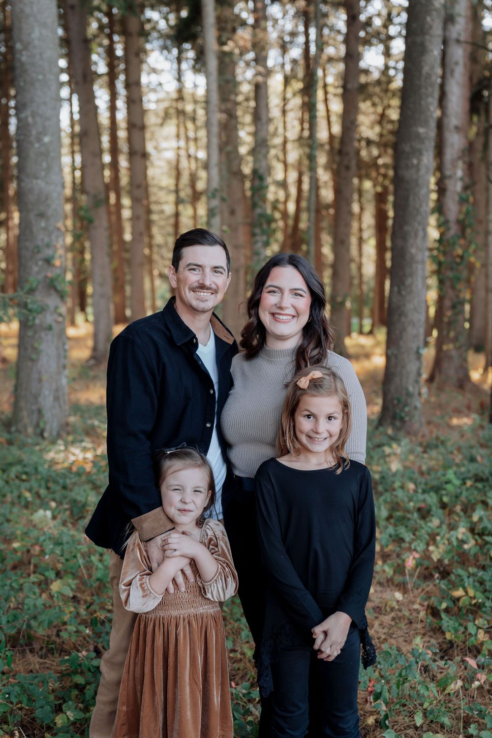 Smiling family of four poses together in pine forest during autumn family photo session.