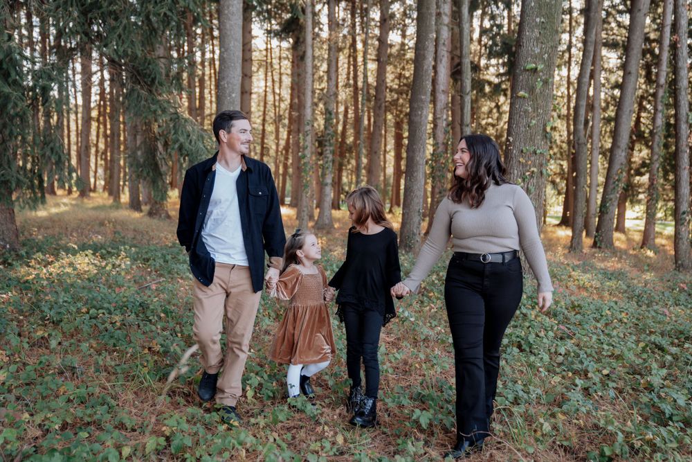 Family of four walks hand-in-hand through sunlit pine forest during fall photo session.