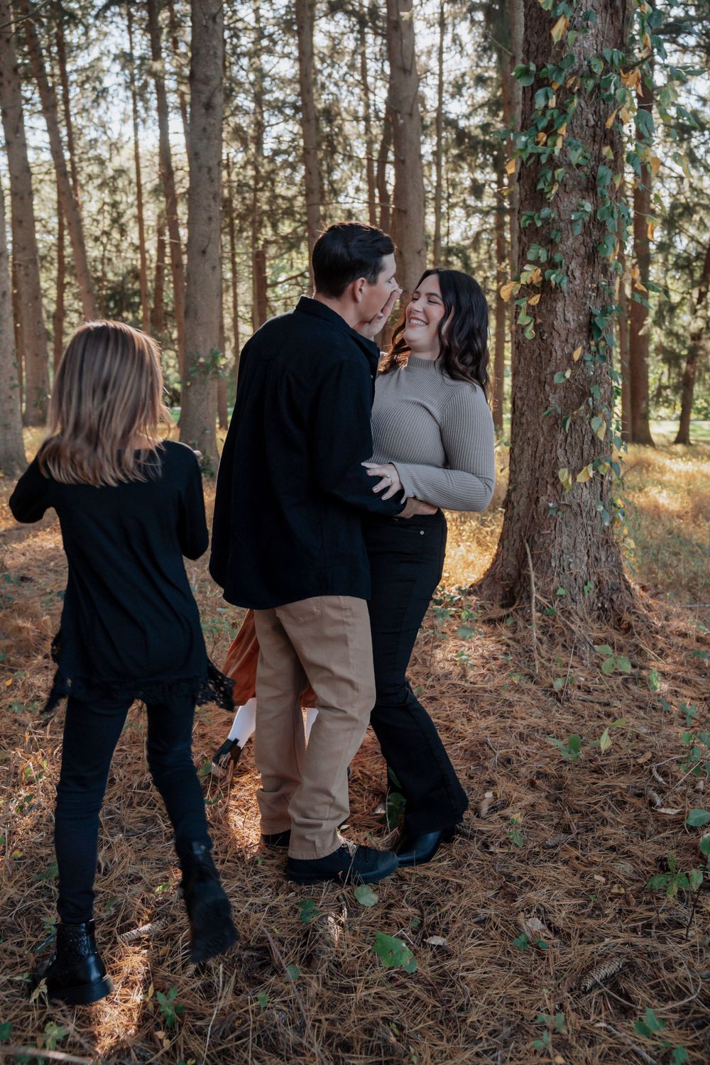 Couple embraces and laughs in pine forest while children play nearby in autumn light.