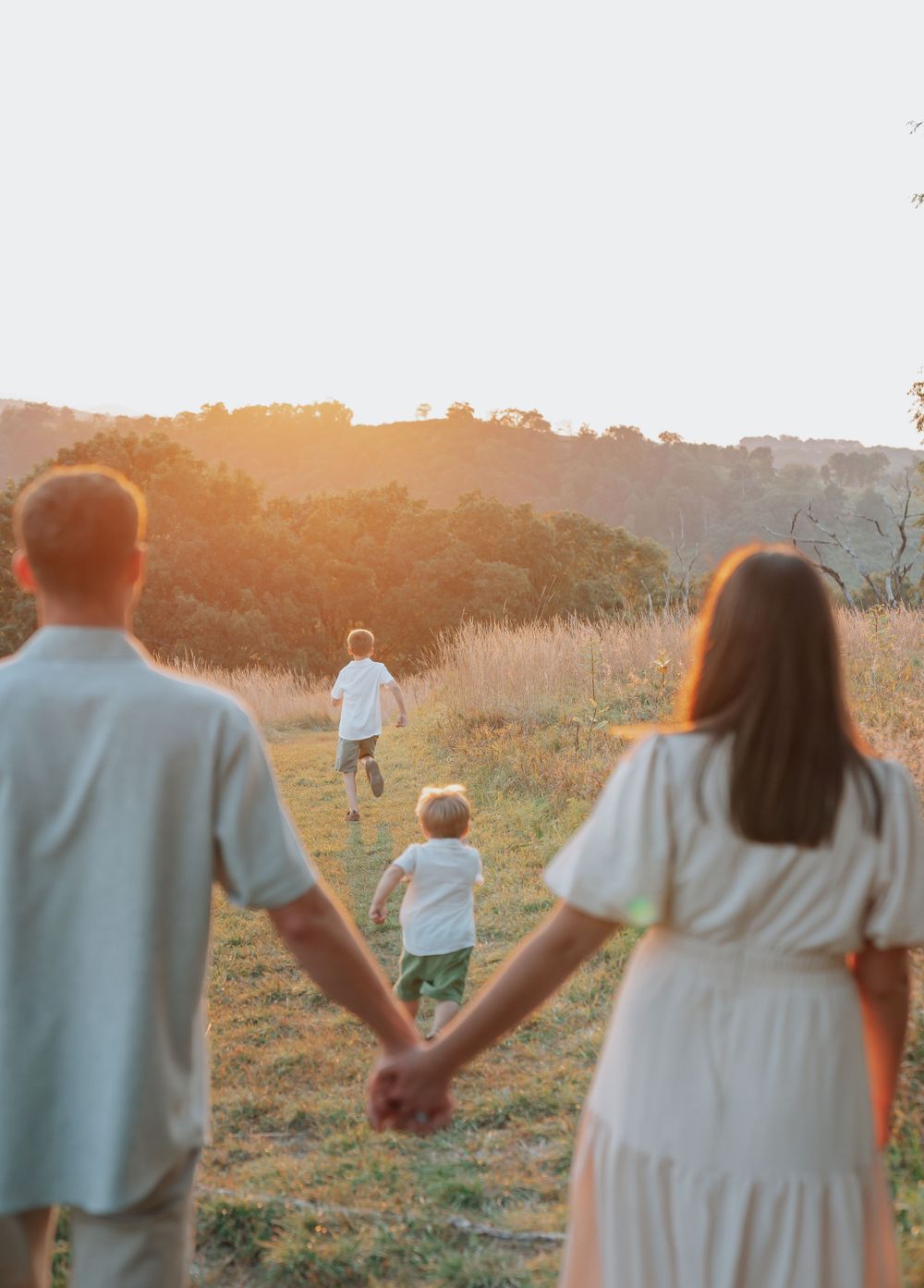 Parents hold hands watching two young children run through a sunlit meadow at golden hour.