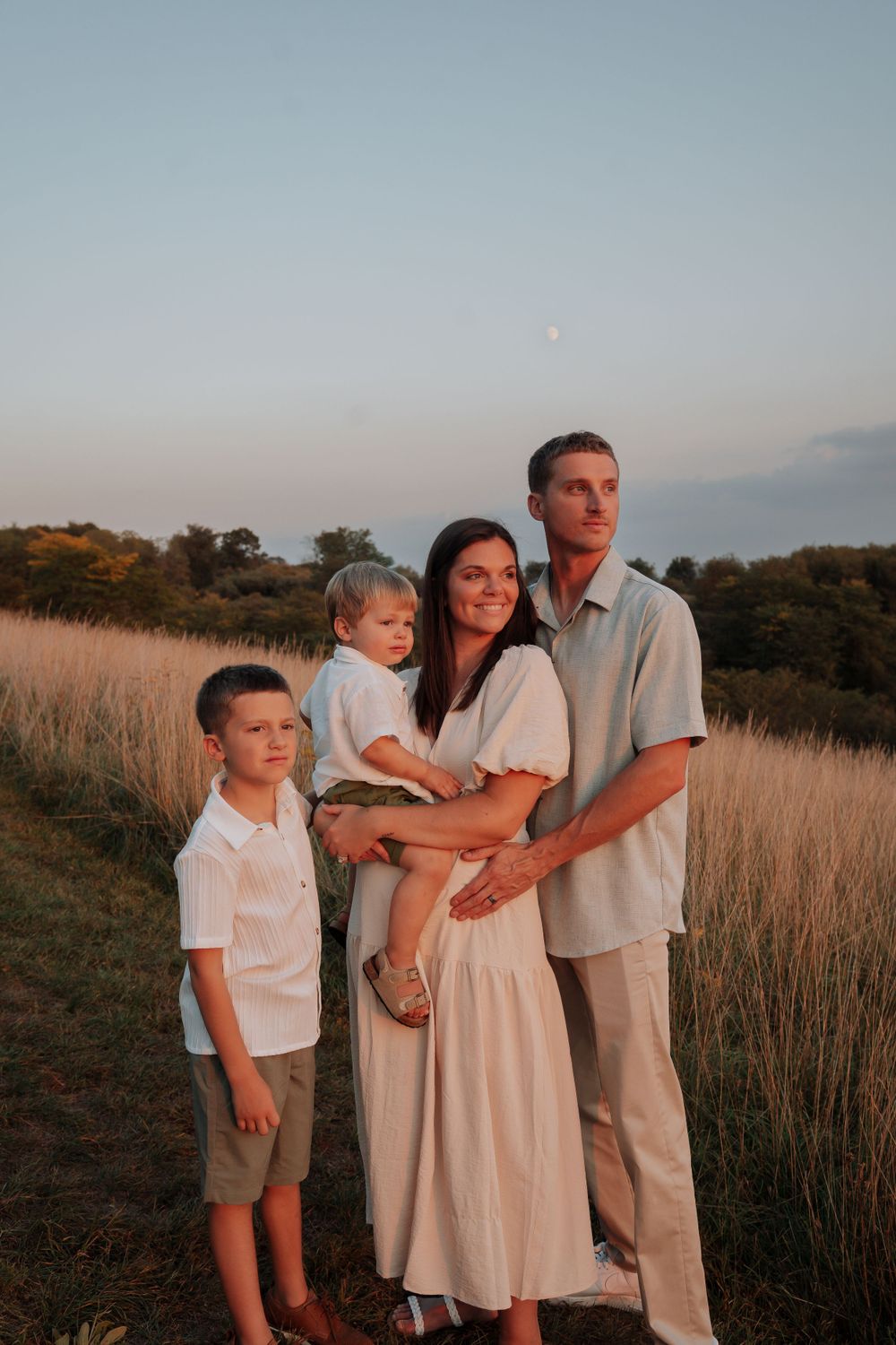 Family of four stands together in a wheat field at dusk with a crescent moon above.