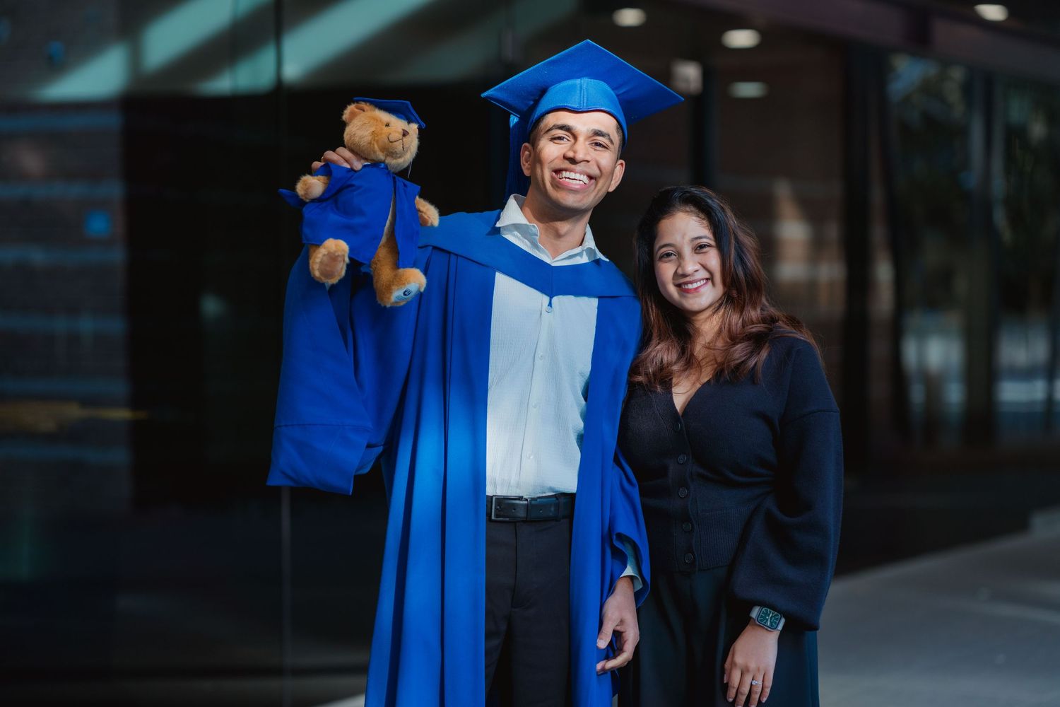 Two college graduates in blue caps and gowns hold hands and smile back at the camera while standing on a sunny, tree-lined lawn.