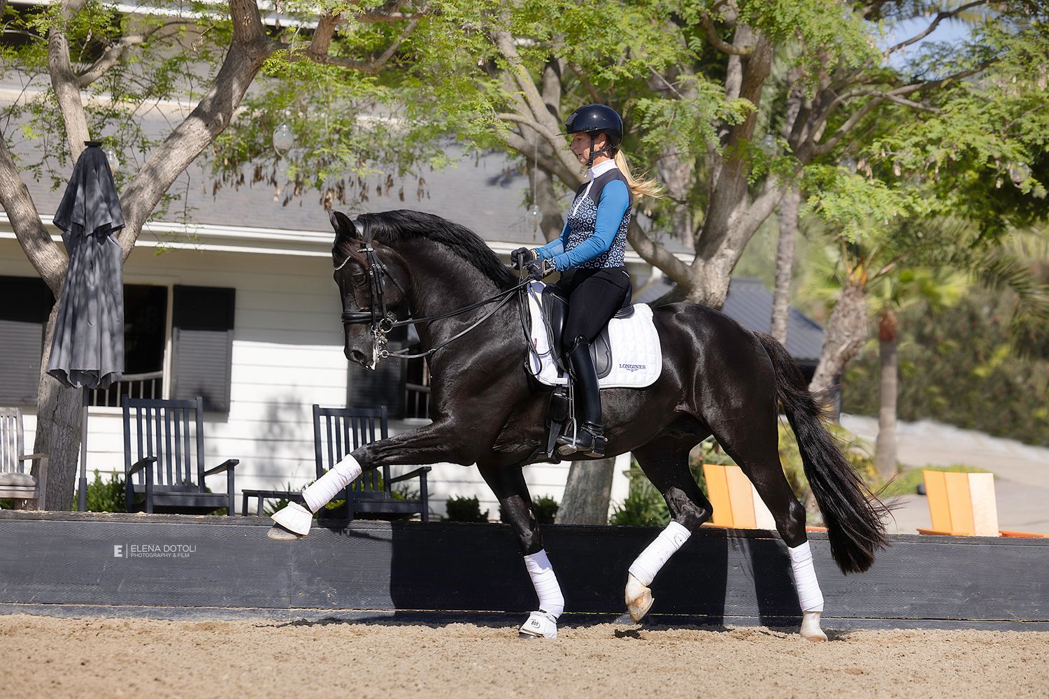 Rider in a black helmet and dark riding attire guides a black horse across a sandy dressage arena beside a white fence and trees.