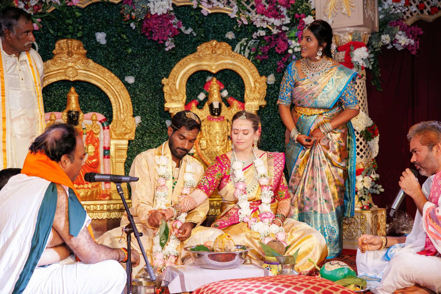 Bride and groom in traditional attire sit before a decorated deity altar during a Hindu wedding ceremony as a priest and guests look on.