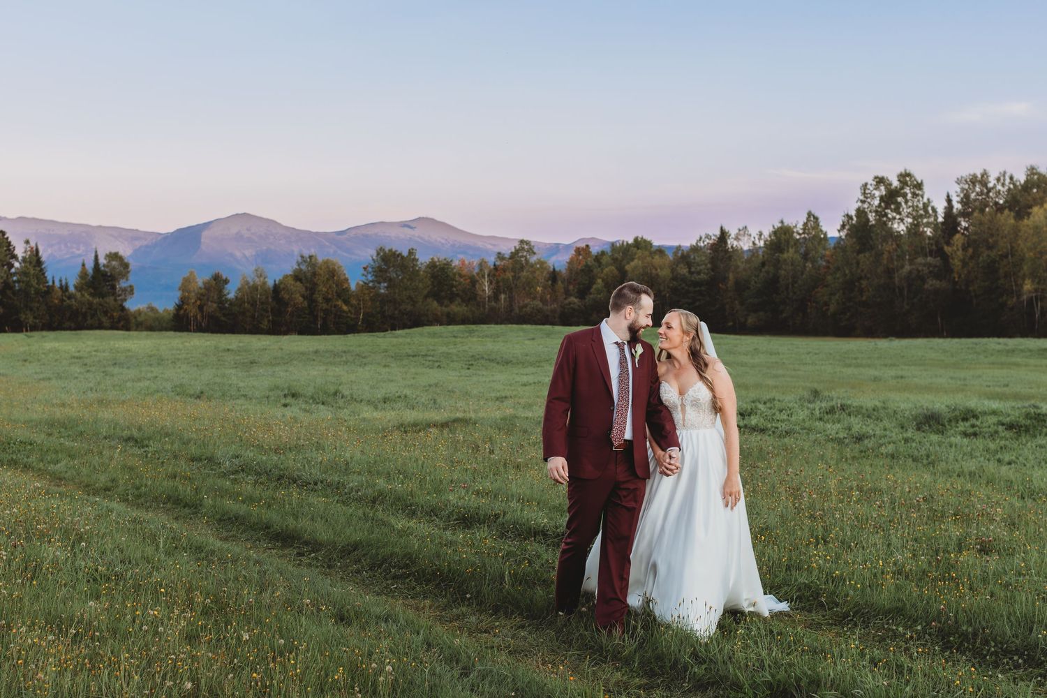 Bride and groom standing together in a green field with mountains beneath a pastel evening sky.