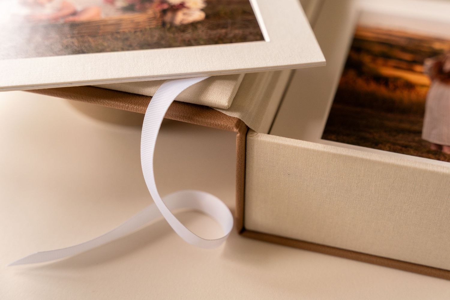 Close-up of an open linen-covered keepsake photo album box with a white satin ribbon and a framed print resting on top.