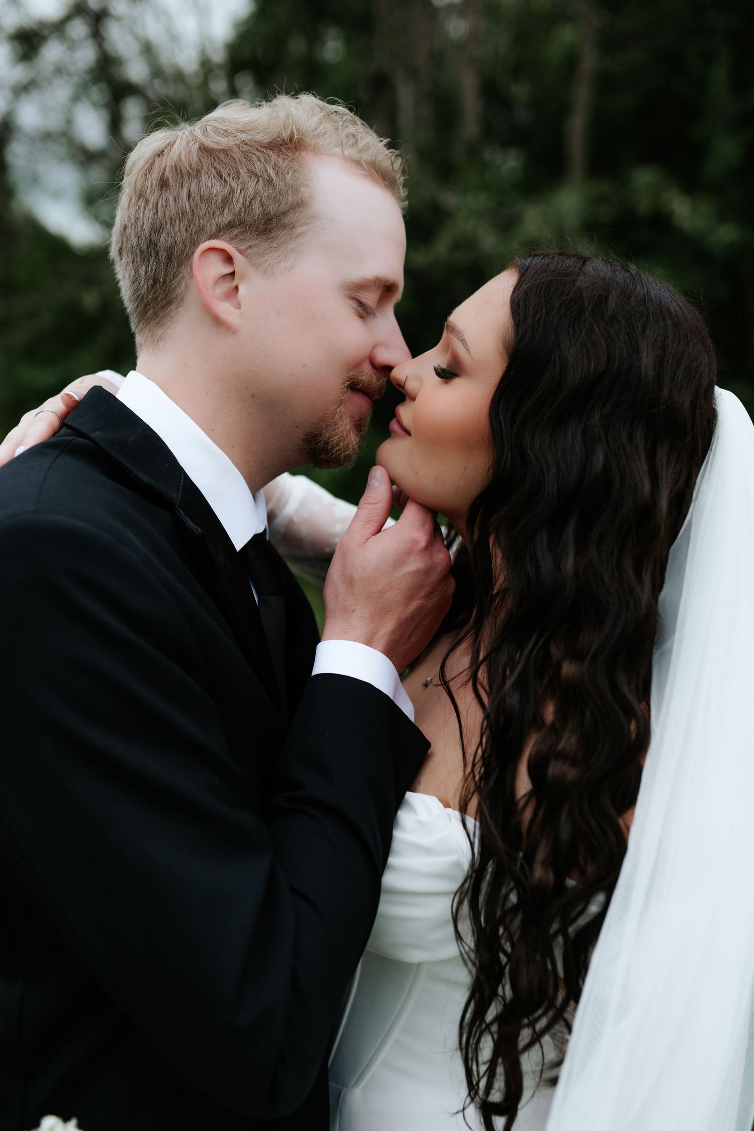 Bride in a white gown and long veil shares an intimate almost-kiss with her groom in a black suit, with soft green trees behind them.