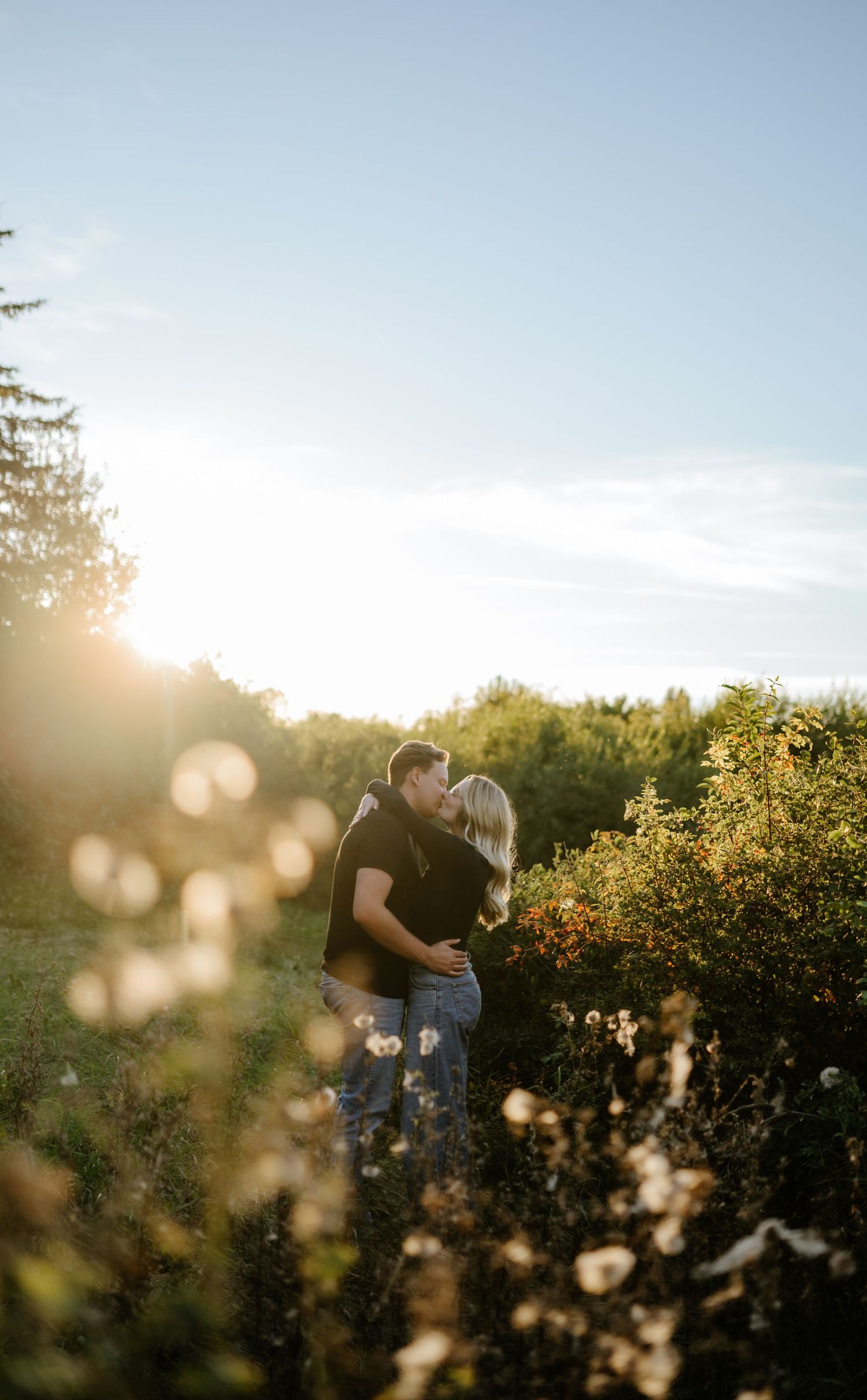 Couple embracing in a sunlit meadow of wildflowers and tall grasses at golden hour, with glowing sun flare and trees in the background.