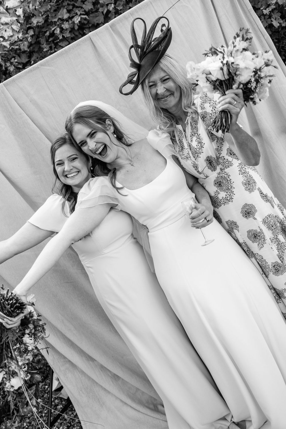 Black and white photo of a laughing bride in a fitted gown posing with two women, one in a fascinator holding a bouquet, at a wedding.