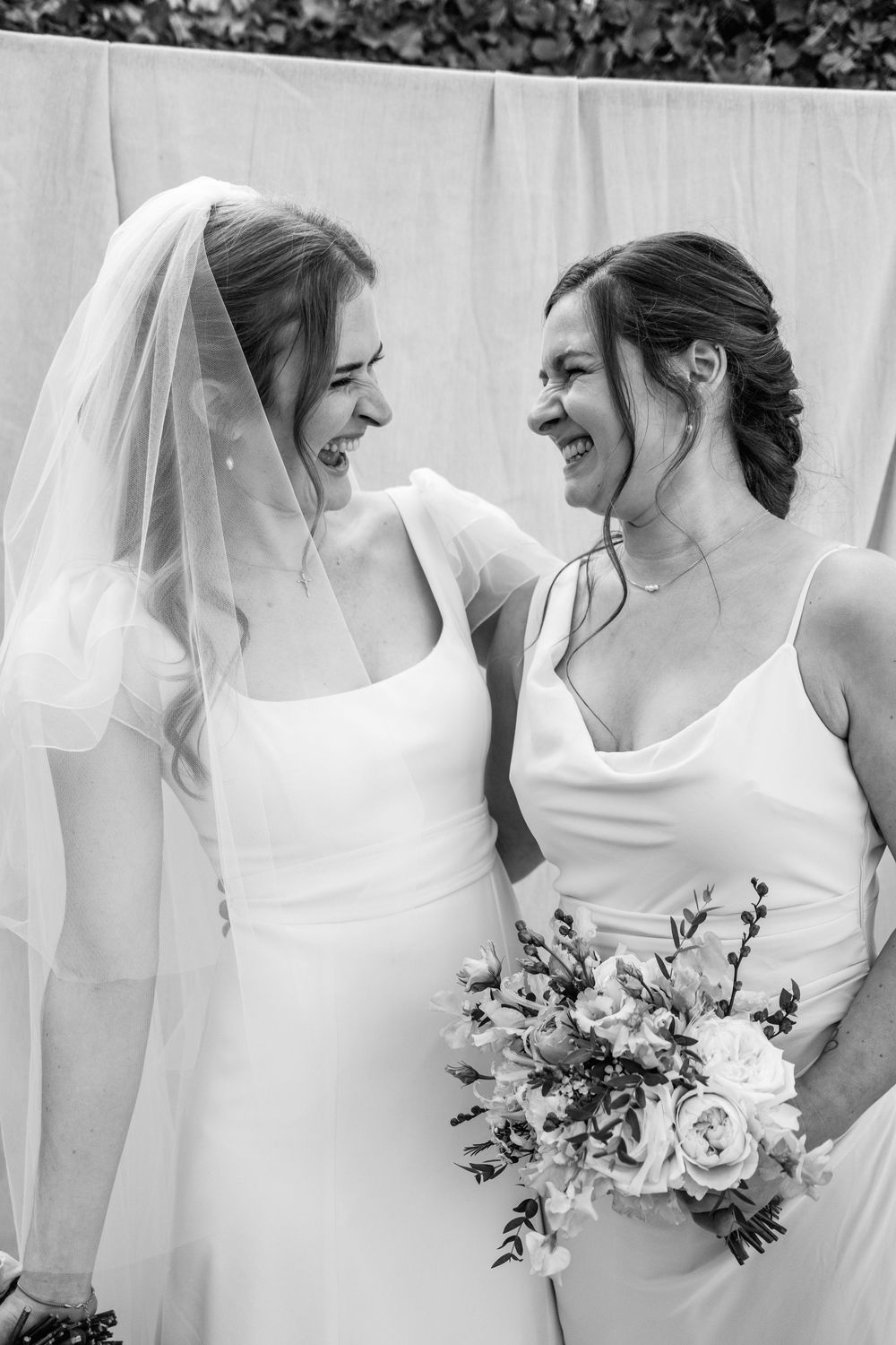 Black and white photo of two women in white dresses laughing together, one wearing a bridal veil and holding a floral bouquet.