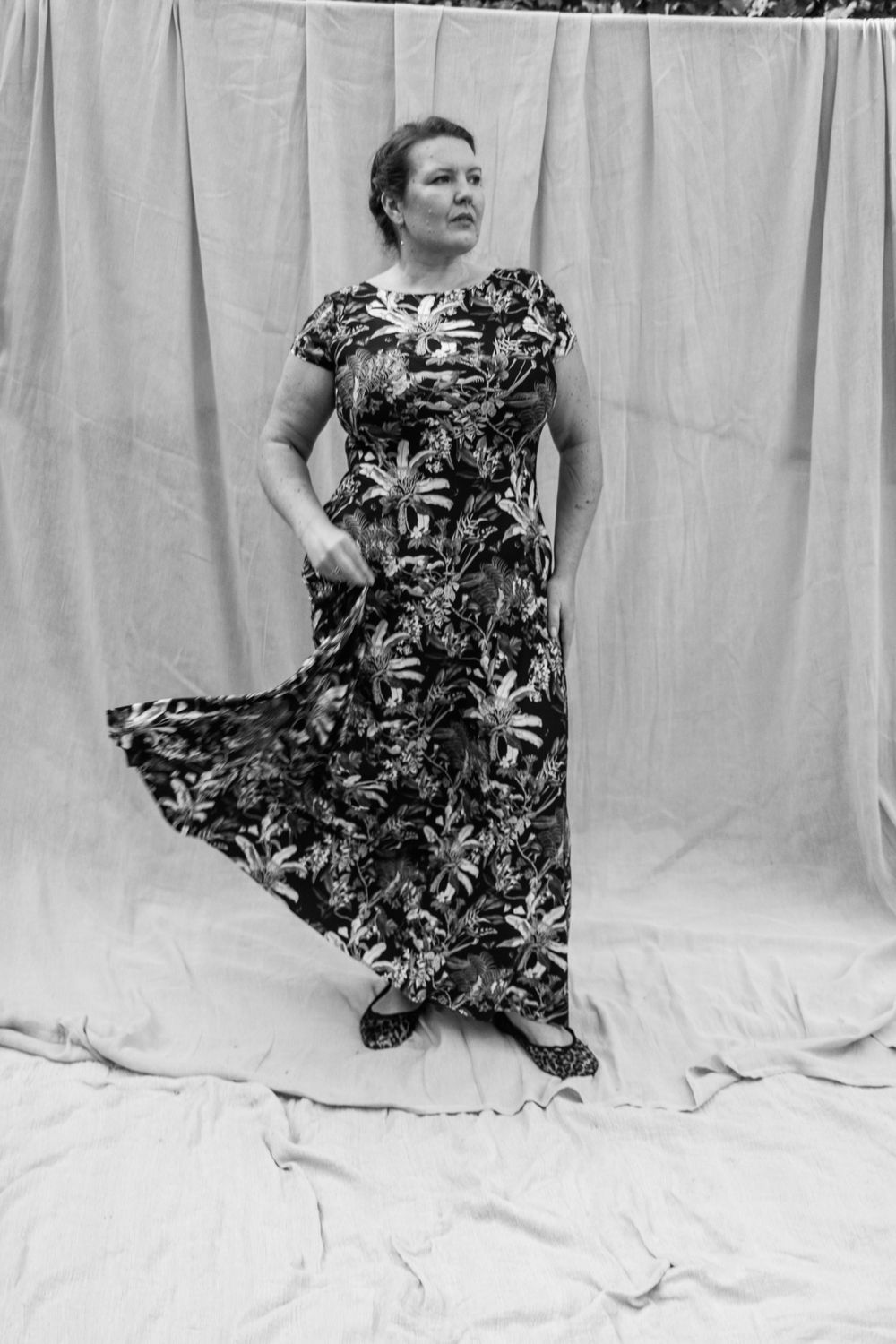 Black and white photo of a woman in a floral maxi dress holding out her skirt while posing against a draped fabric backdrop.