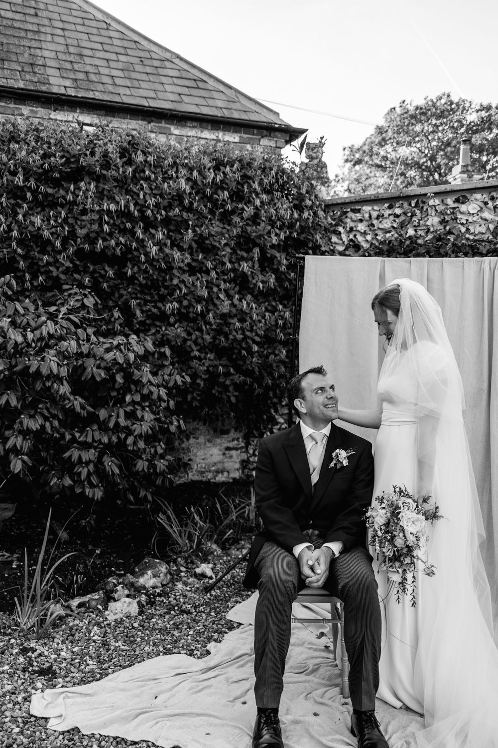 Black and white photo of a bride in a veil holding a bouquet, touching a seated groom's shoulder in a garden by an ivy hedge.