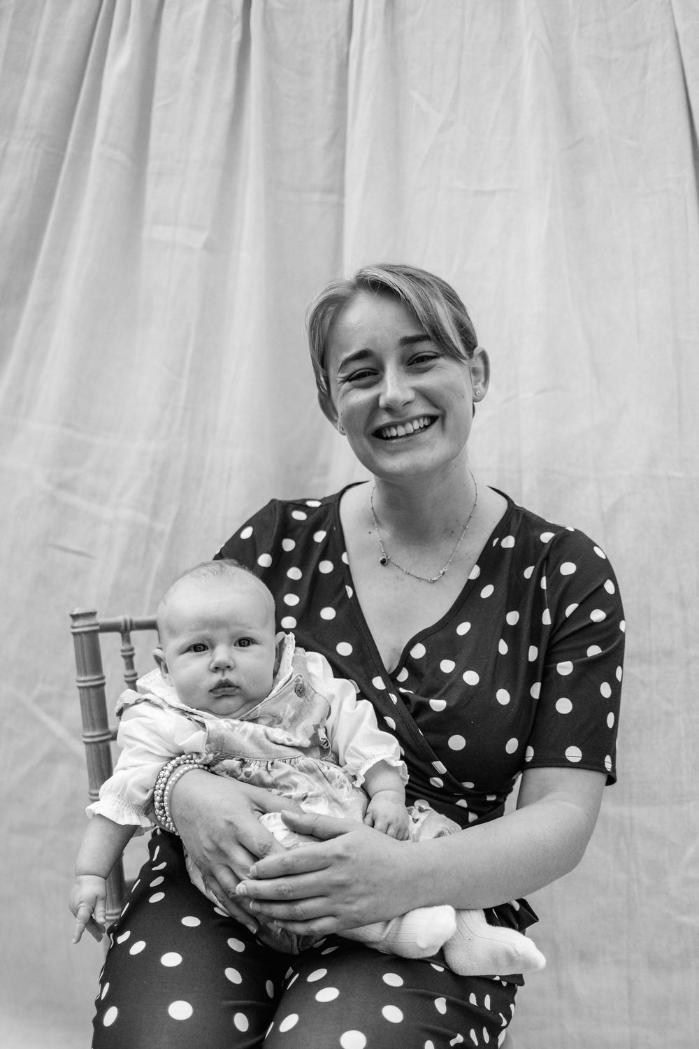 Smiling woman in a polka dot dress sits on a chair holding a baby in a black and white portrait against a draped backdrop.