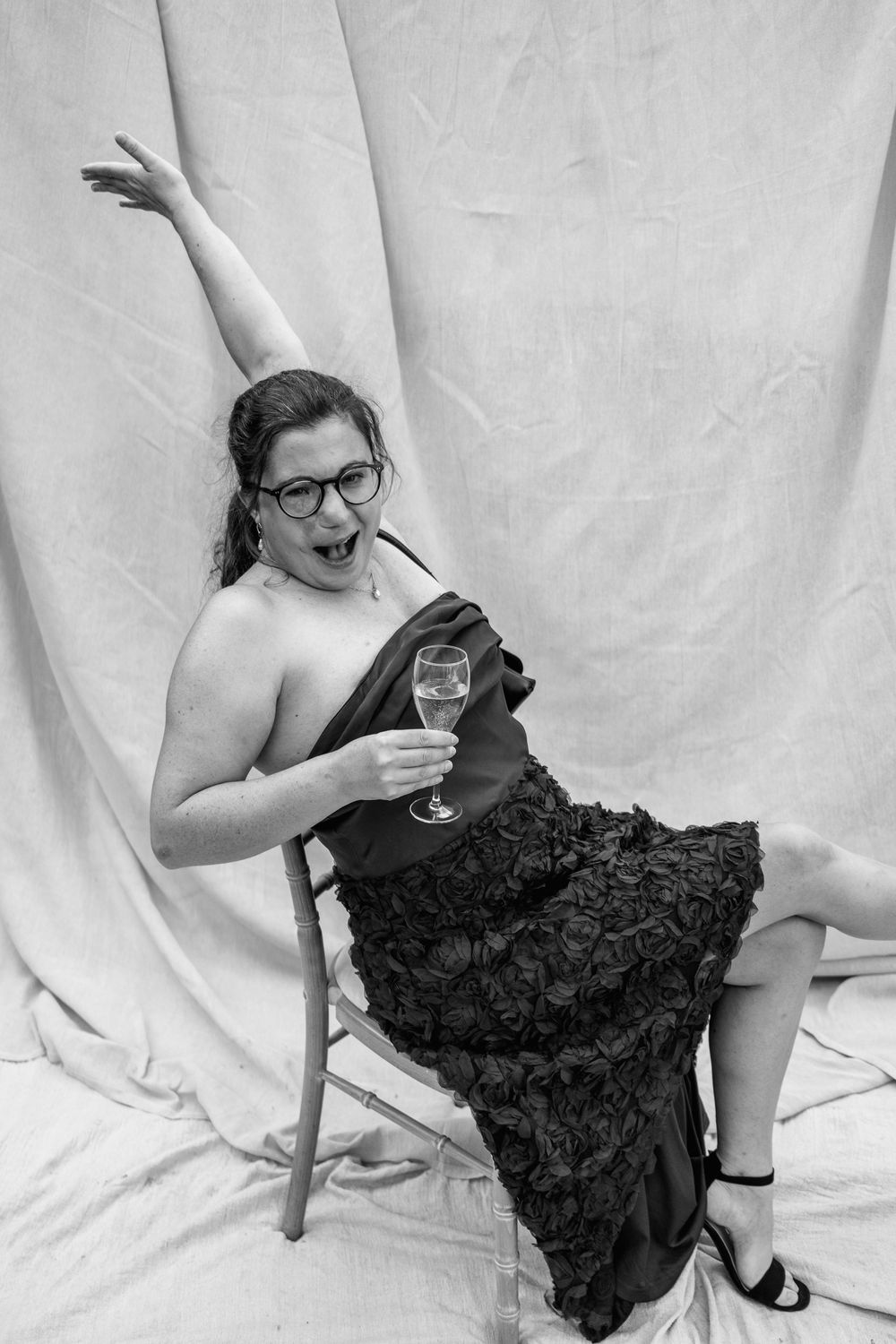 Playful woman in glasses and a ruffled dress raises one arm and holds a champagne glass while seated in a black and white portrait.
