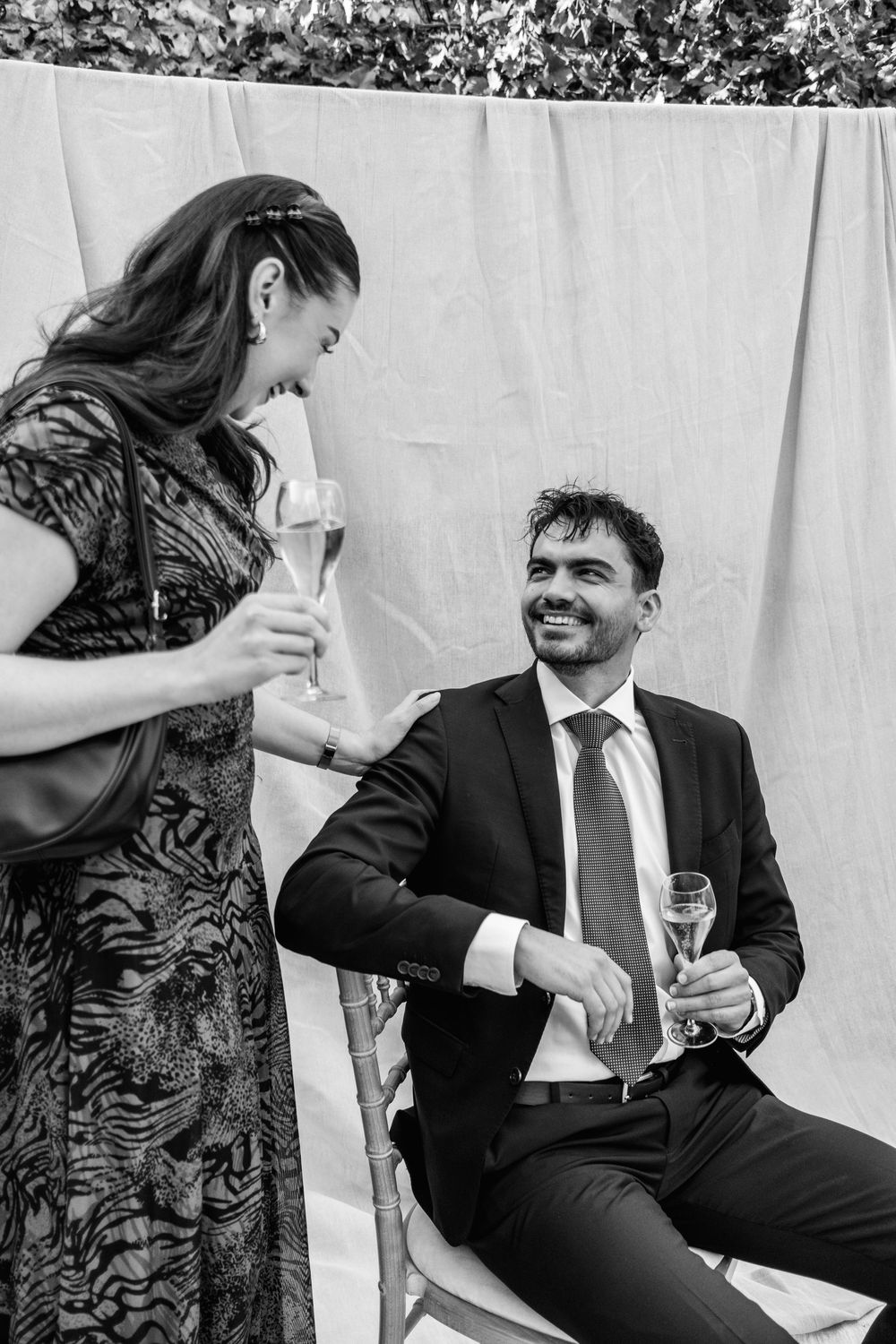 A smiling woman in a patterned dress touches the shoulder of a seated man in a suit as they hold champagne glasses at a wedding.