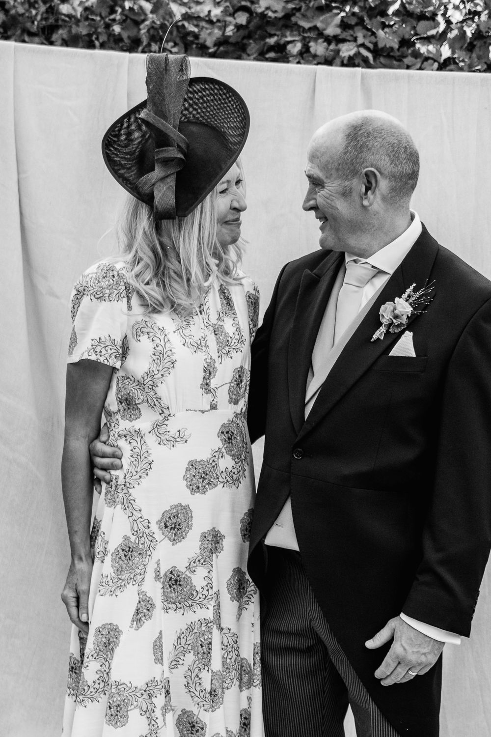 Black and white photo of a smiling couple at a wedding, she in a floral dress and fascinator, he in a morning suit with a boutonniere.