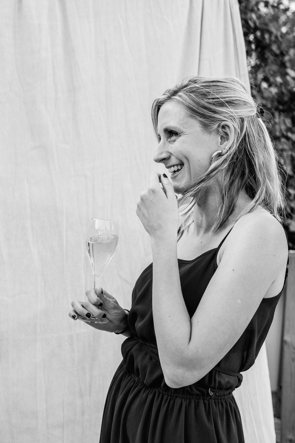 Black and white photo of a smiling woman in a sleeveless dress holding a champagne flute against a draped white backdrop.