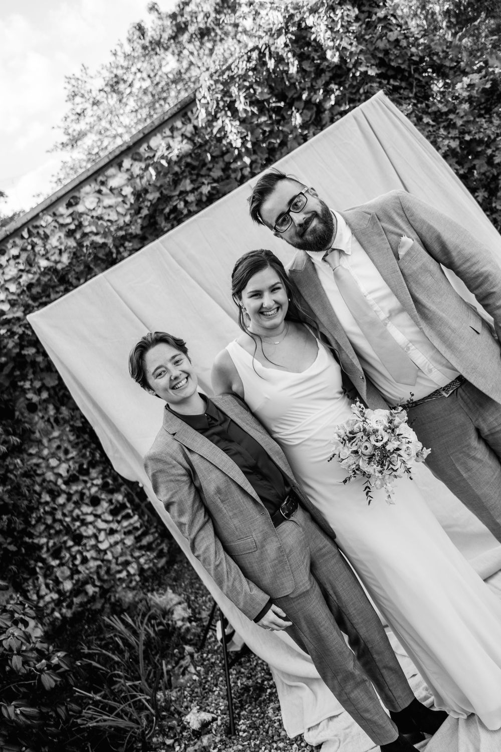 Black and white wedding photo of a smiling bride holding a bouquet, posing with two people in suits before a white backdrop and ivy wall.
