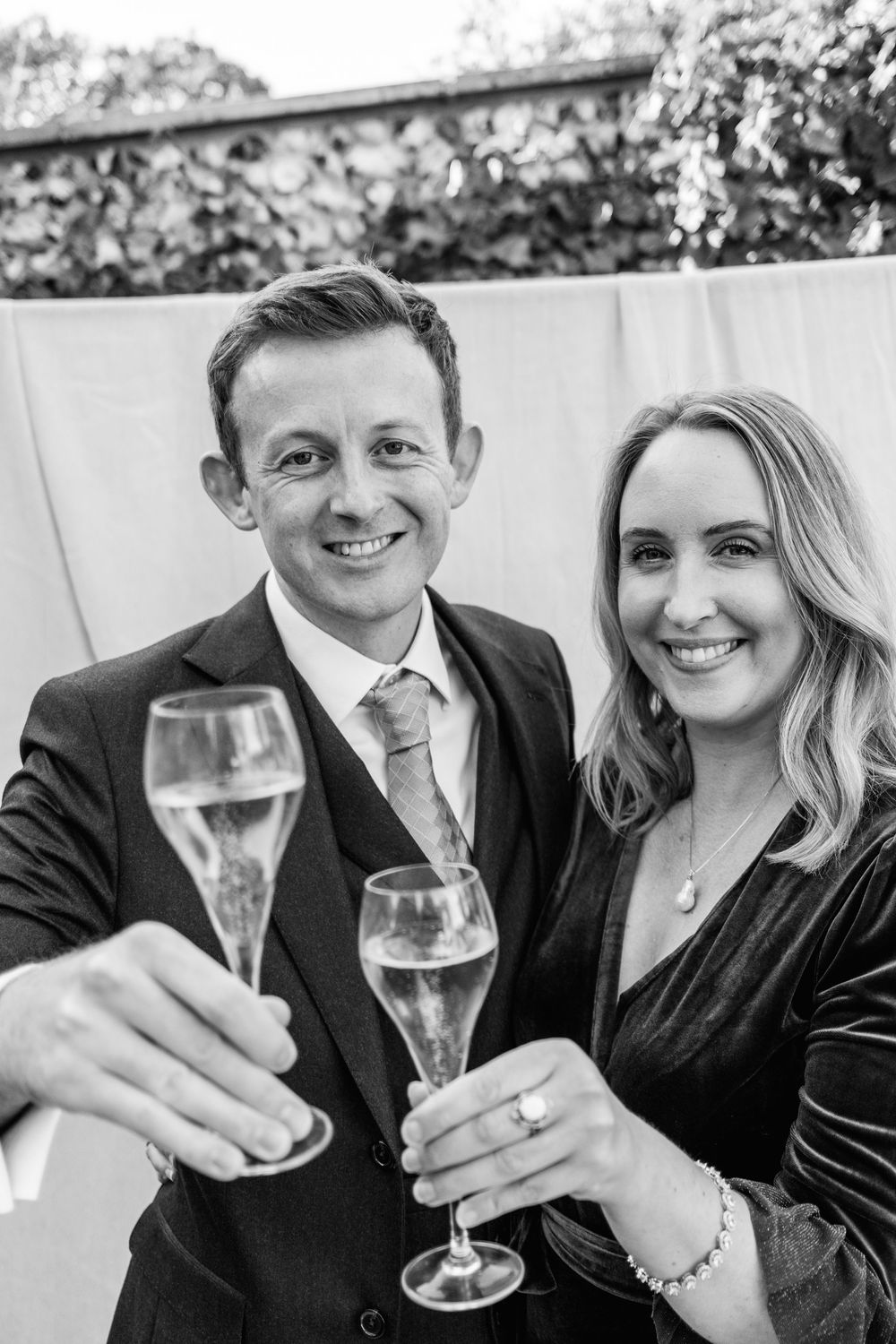 Smiling man in a suit and woman in a velvet dress toast with champagne flutes in a black and white celebration portrait.