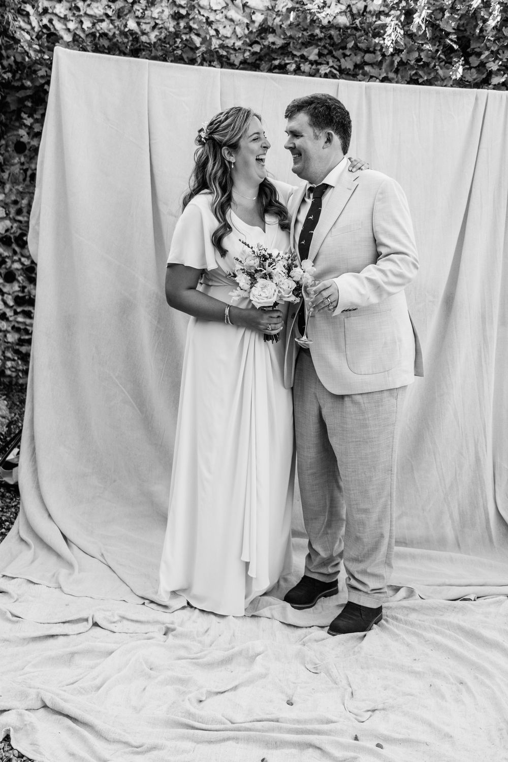 A smiling bride in a flowing white dress holding a bouquet laughs with her groom in a light suit in front of a draped backdrop.