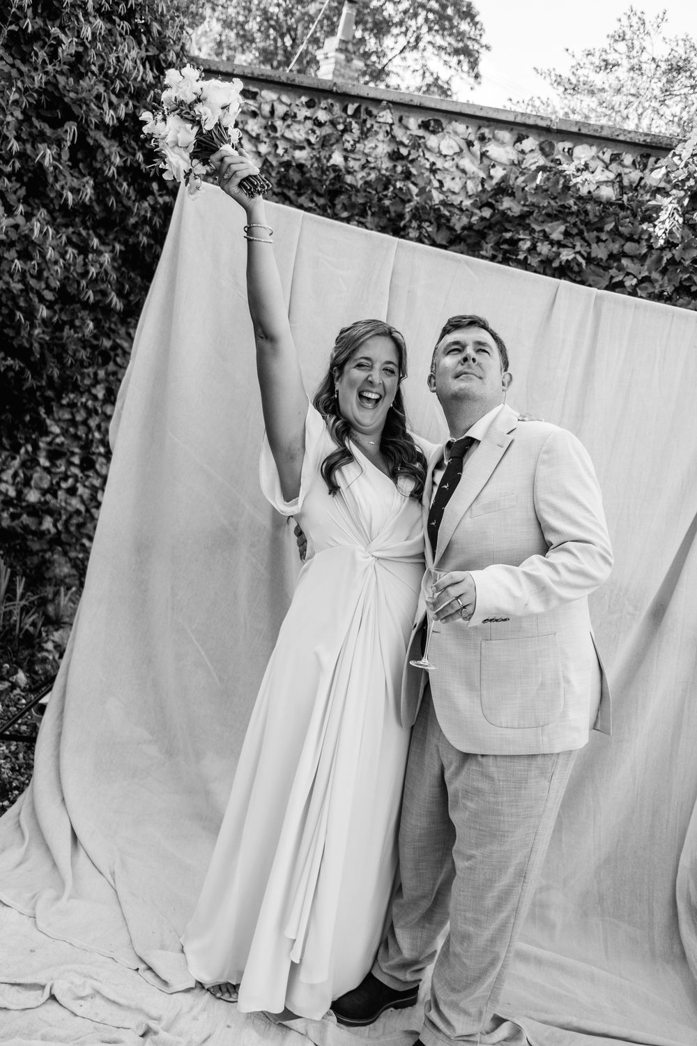 A joyful bride raises her bouquet in the air while laughing beside her groom in a light suit in front of a draped fabric backdrop.