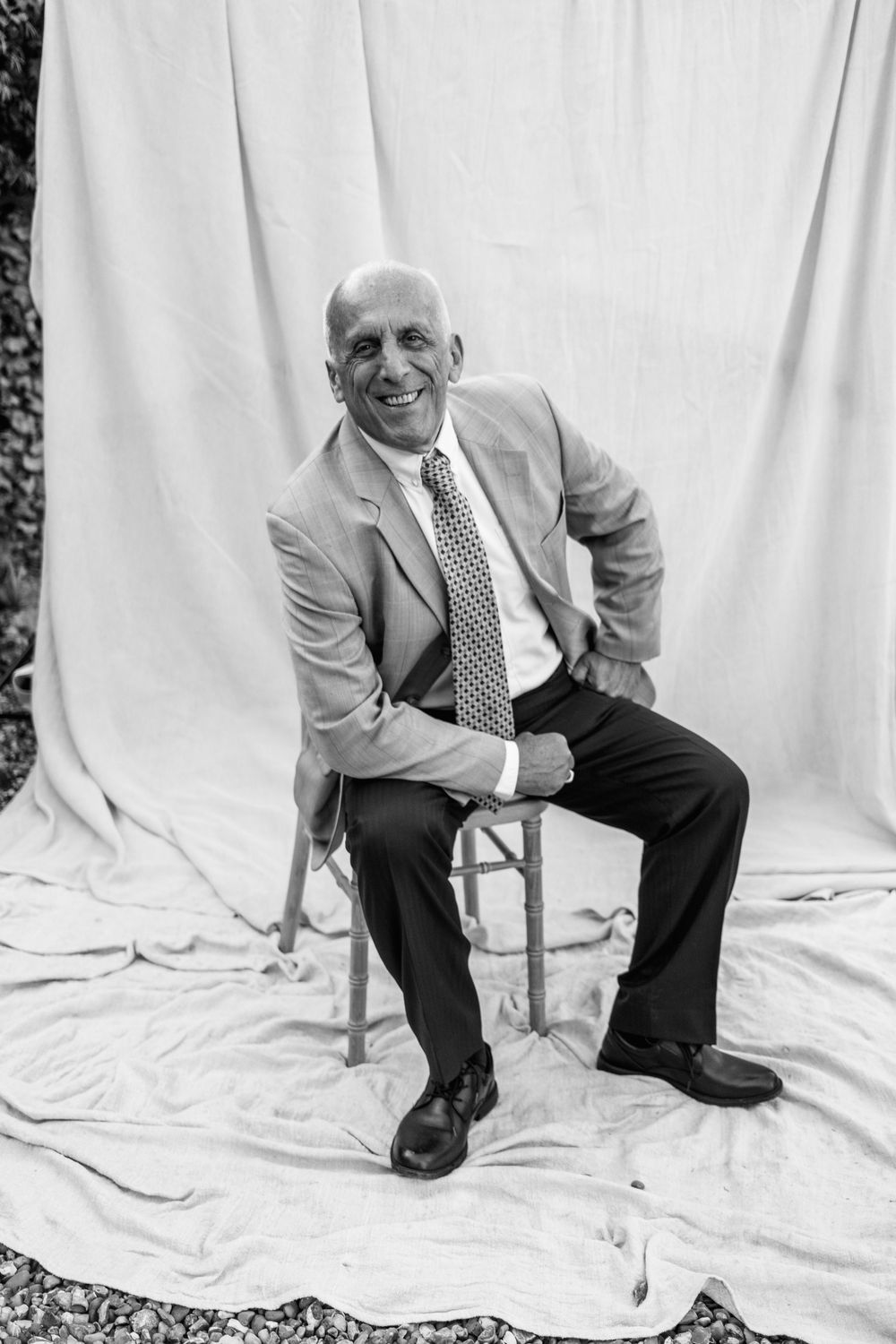 Black and white portrait of a smiling bald man in a blazer and patterned tie, seated on a wooden stool against a draped fabric backdrop.