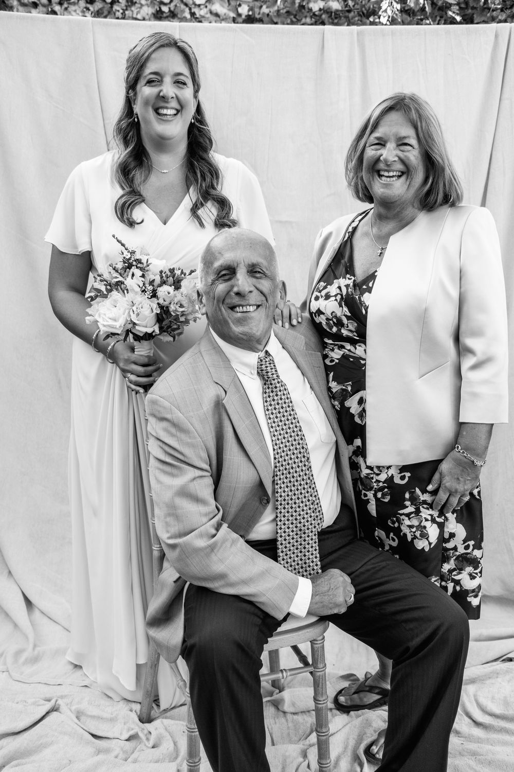Black and white wedding portrait of a smiling bride holding a bouquet with a seated man in a suit and a woman in a floral dress.
