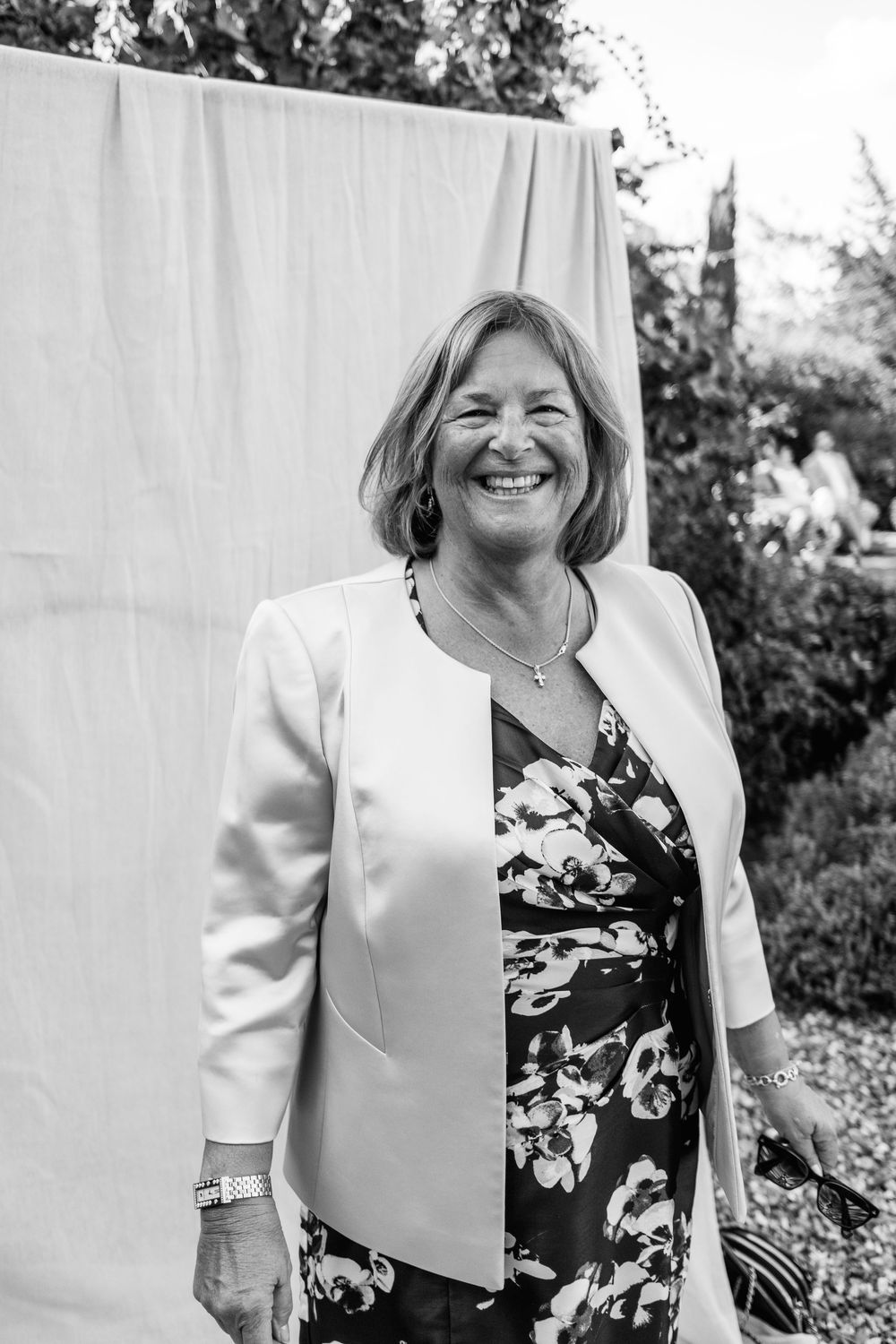 Black and white portrait of a smiling woman in a light blazer and floral dress standing outdoors in front of a hanging white sheet.