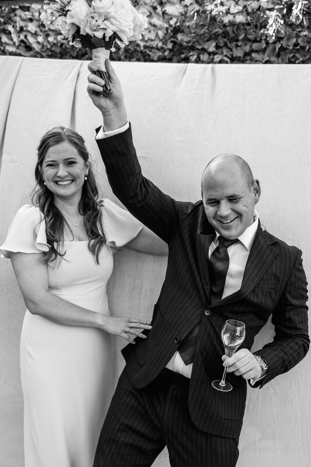 Black and white wedding photo of a smiling bride in a white dress beside a groom in a pinstripe suit raising a bouquet and holding champagne