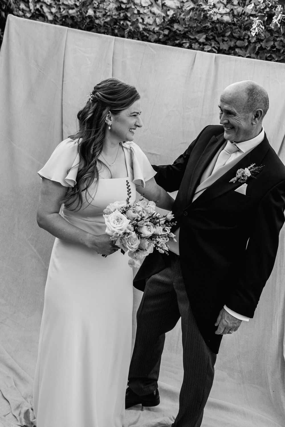 Black and white wedding photo of a smiling bride holding a bouquet laughing with a man in a morning suit and boutonniere.