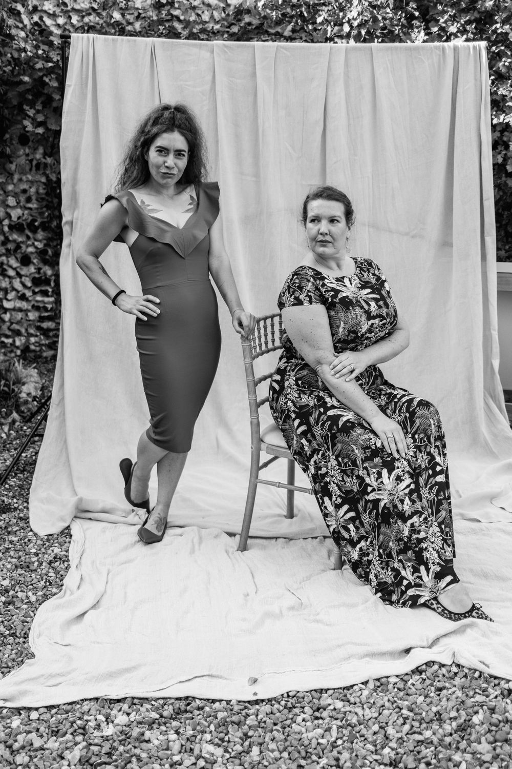 Black and white portrait of two women posing against a draped backdrop, one standing in a fitted dress and one seated in a floral gown.