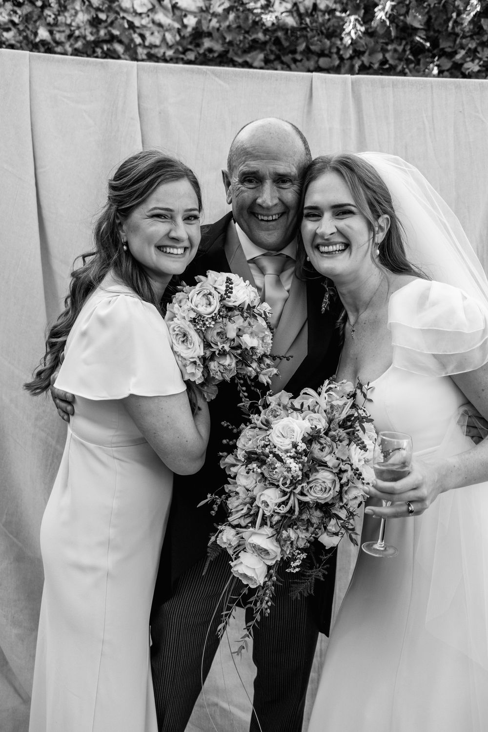 Black and white wedding portrait of a smiling bride and bridesmaid embracing a grinning man in a suit, both holding floral bouquets.