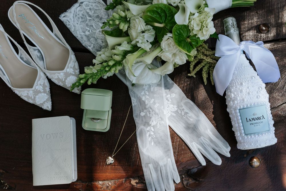Bridal flat lay with white lace heels, sheer gloves, a green and white bouquet, ring box, vow book and a ribboned champagne bottle on wood.