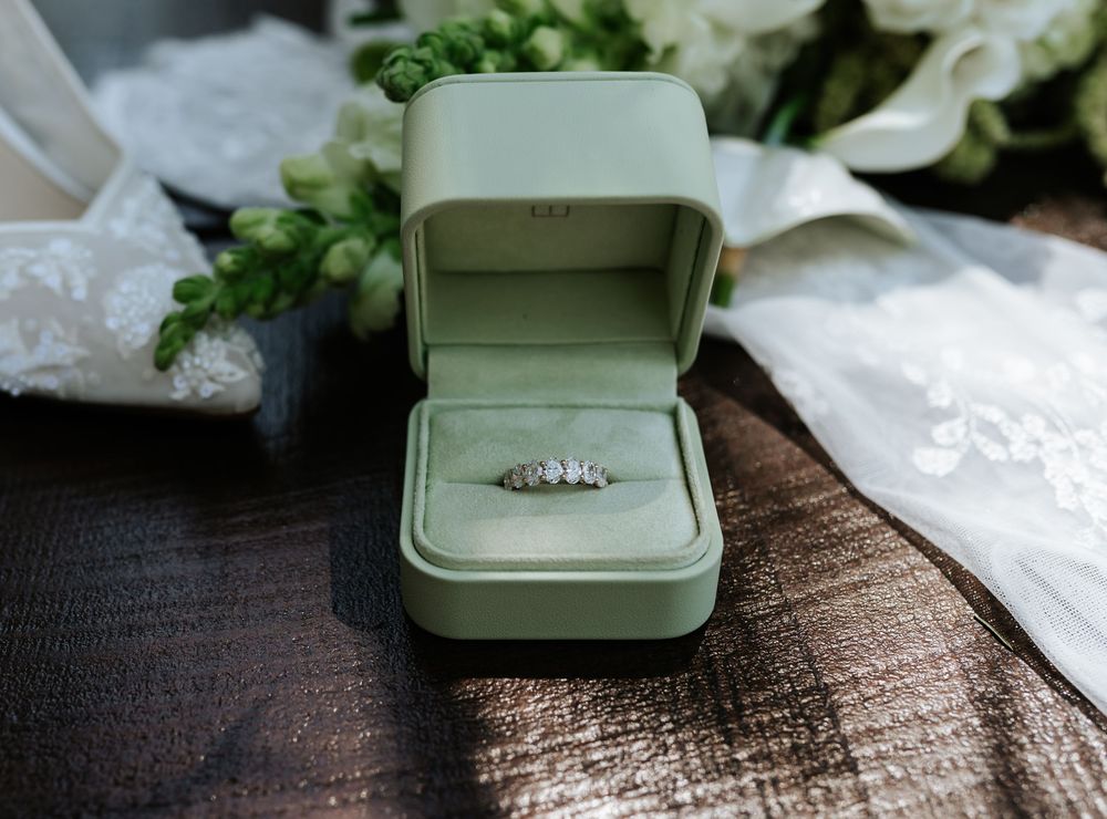 Diamond engagement ring in an open sage green ring box on a dark wooden table beside white lace fabric and fresh white flowers.