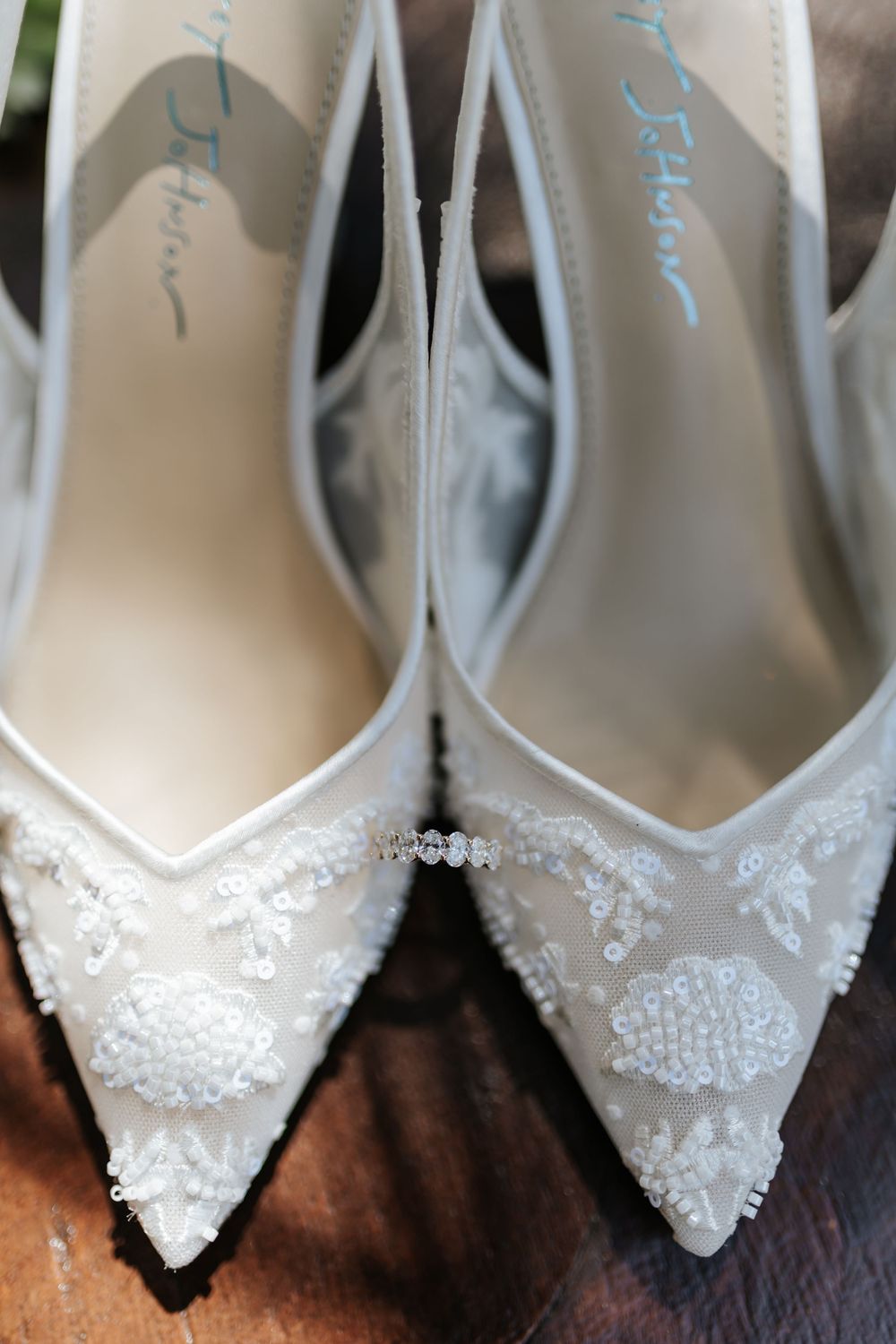 Pair of white pointed-toe bridal heels with beaded lace appliqué, a crystal strap detail, and blue script lettering on the insoles.
