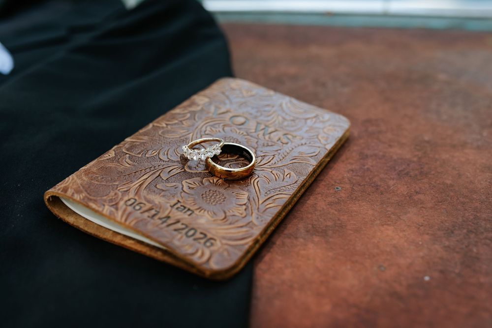 Wedding bands and a diamond engagement ring resting on a tooled brown leather vow book with floral embossing beside a black suit.