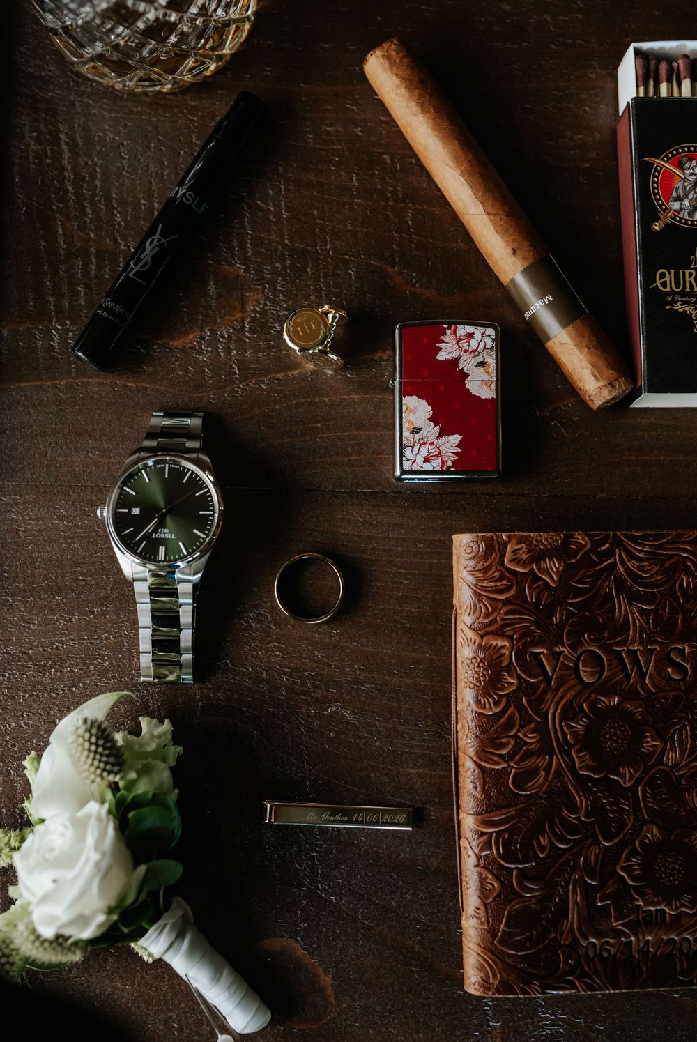 Flat lay of groom's wedding day essentials including a steel wristwatch, cigar, lighter, rings, tie bar, vows book and boutonniere.