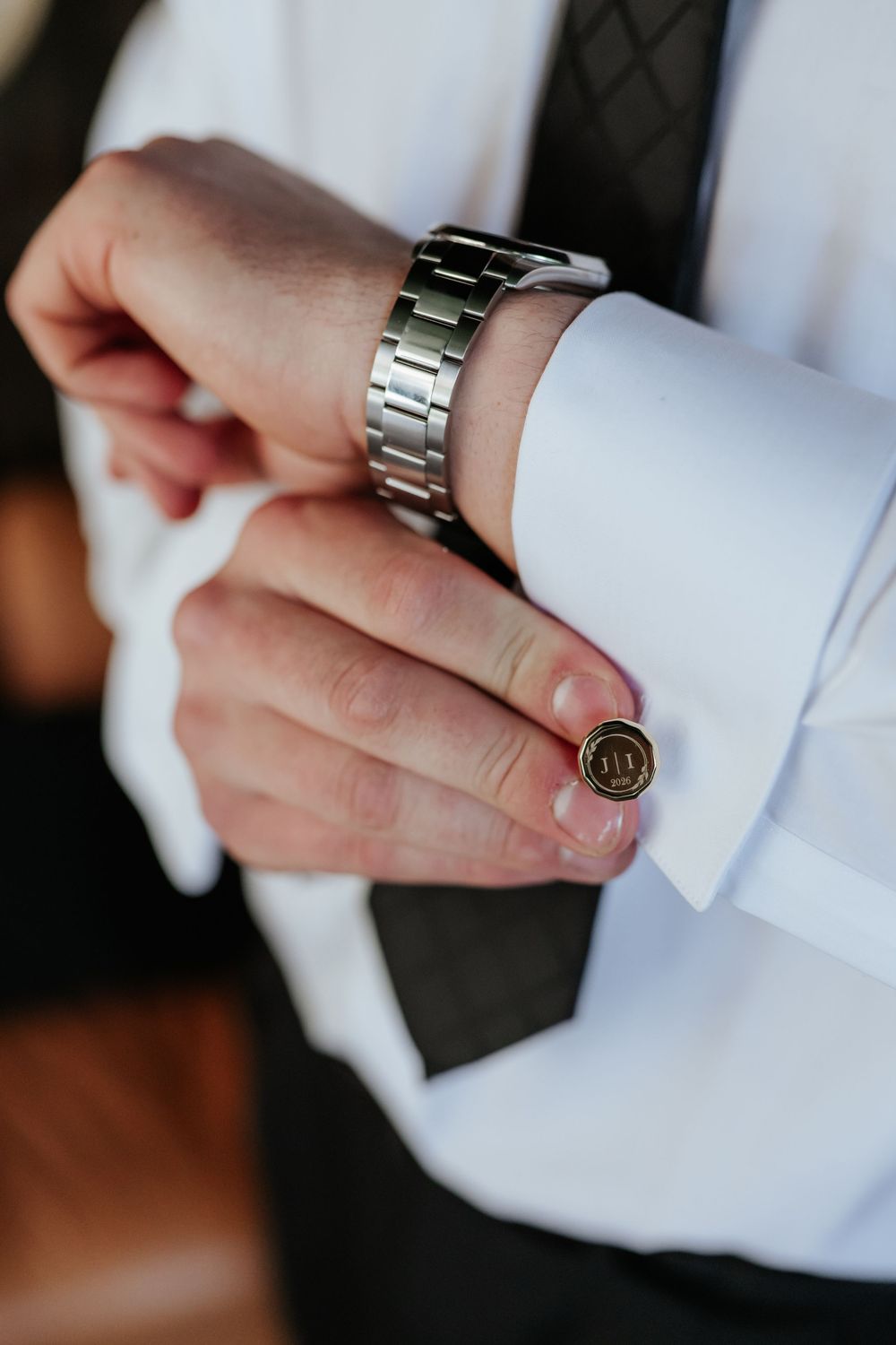 Groom in a white dress shirt and dark tie adjusts a round monogrammed cufflink while wearing a silver stainless steel wristwatch.