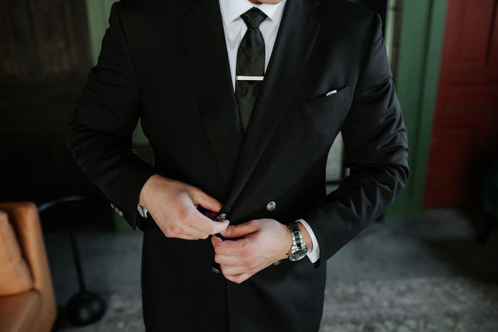 Groom in a black suit and dark tie with tie bar adjusting his jacket button, wearing a wristwatch, in a rustic indoor setting.